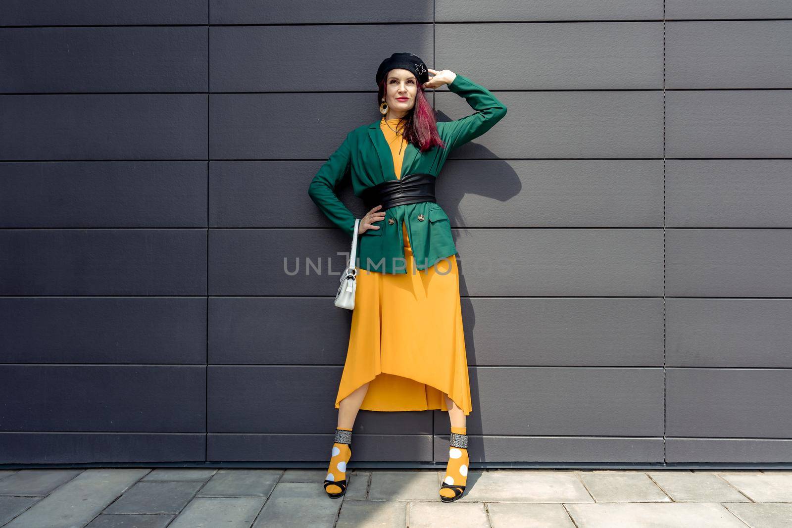 Young woman in bright clothes, yellow skirt and green jacket. Yellow socks in sandals, beret on the head, hair with the color of magenta. Caucasian female fashion model standing against gray wall background, open with copy space