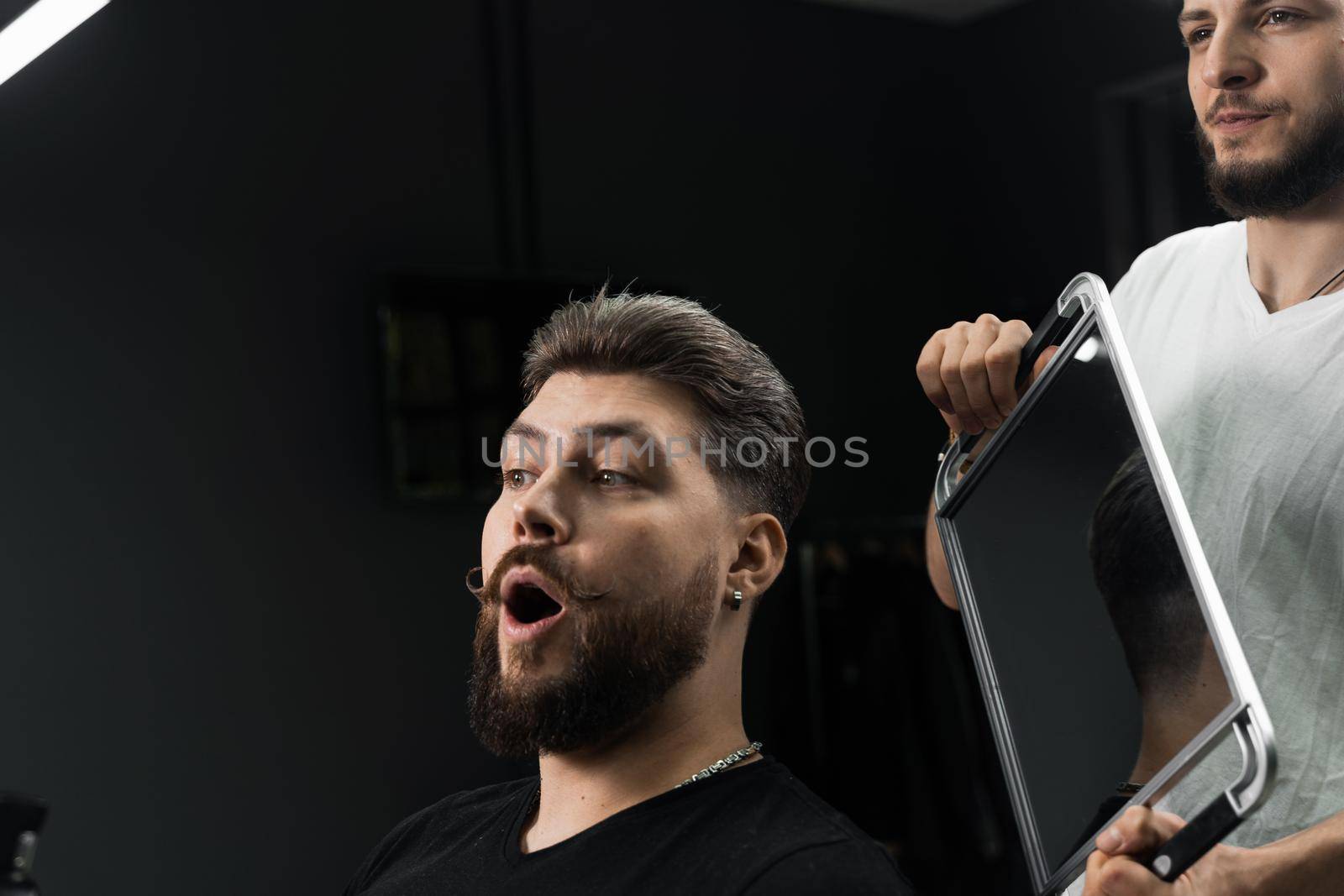 Happy and surprised bearded man looking in mirror in barbershop. Showing result of low fade machine hair.