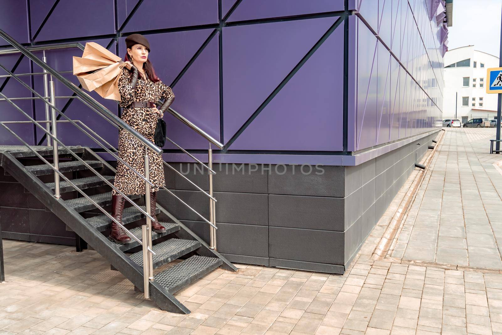 A happy shopaholic girl keeps her bags near the shopping center. A woman near the store is happy with her purchases, holding bags. Dressed in a leopard print dress. Consumer concept. by Matiunina