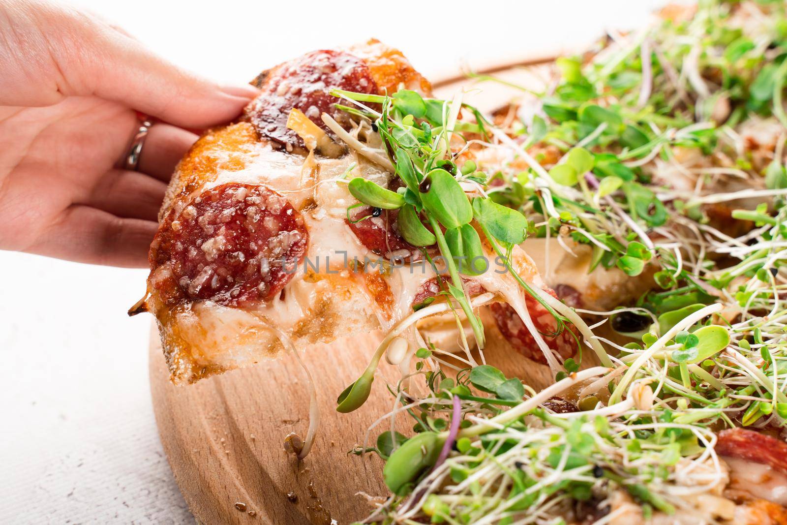 Piece of pinsa romana in hand with stretching mozzarella with salami, cheese, mushrooms, decorated with microgreens on wooden board.