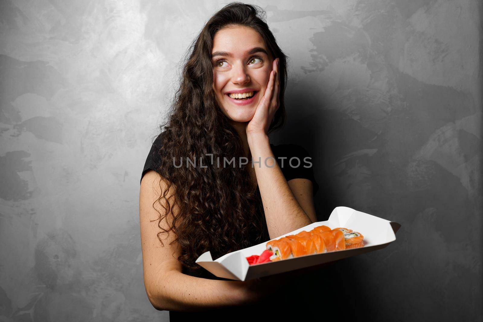 Surprised girl with sushi set philadelphia rolls in a paper box happy girl holding on a gray background. Food delivery