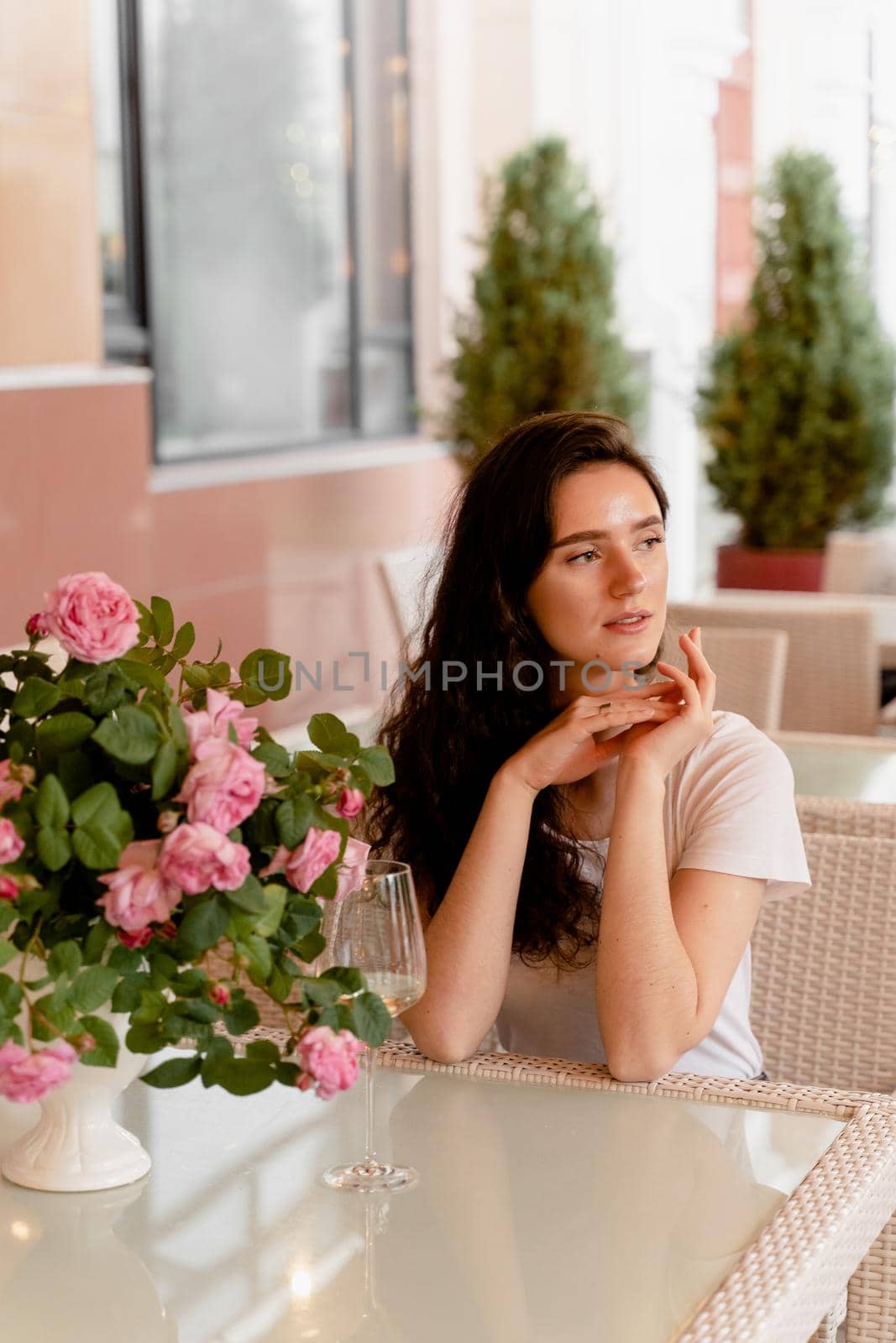 Dreaming woman with glass of alcoholic white wine in cafe in summer terrace and bouquet of roses on the table.
