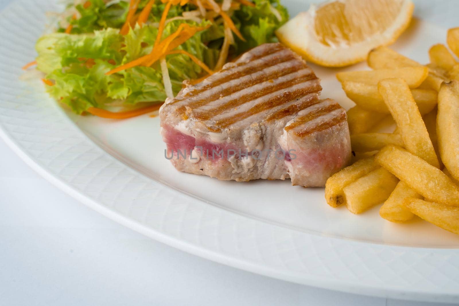 Tuna steak grilled with french fries salad of carrots, parsnips, lettuce and lemon on white plate on white background