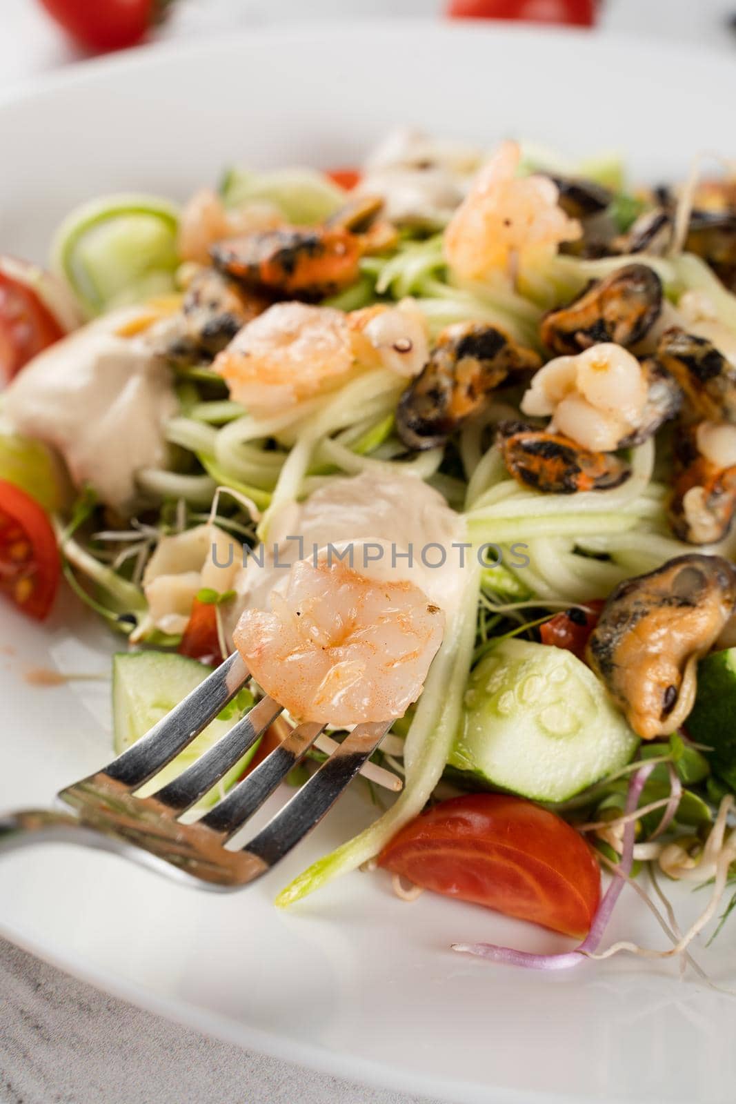 Salad with mussel, shrimp, tomato cherry, cucumber, zucchini pasta, microgreen, yogurt sauce on white plate on light table