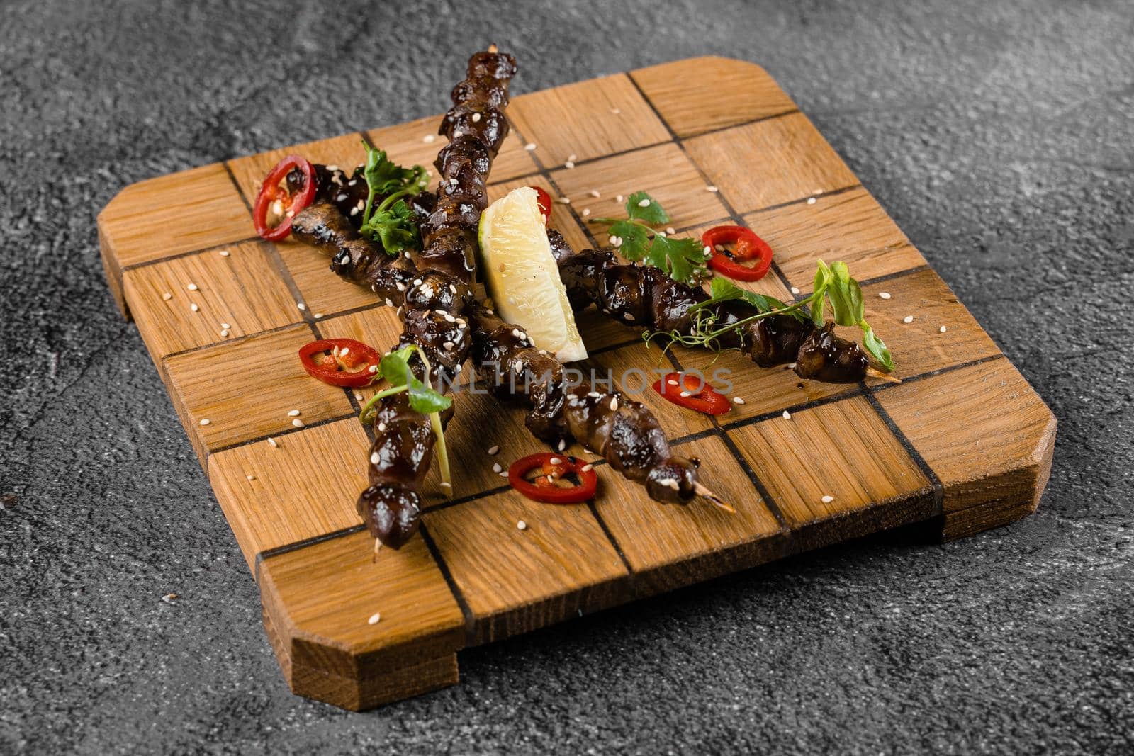 Grilled chicken hearts with lime on wooden skewer, garnished with chili, cilantro, sesame on gray background