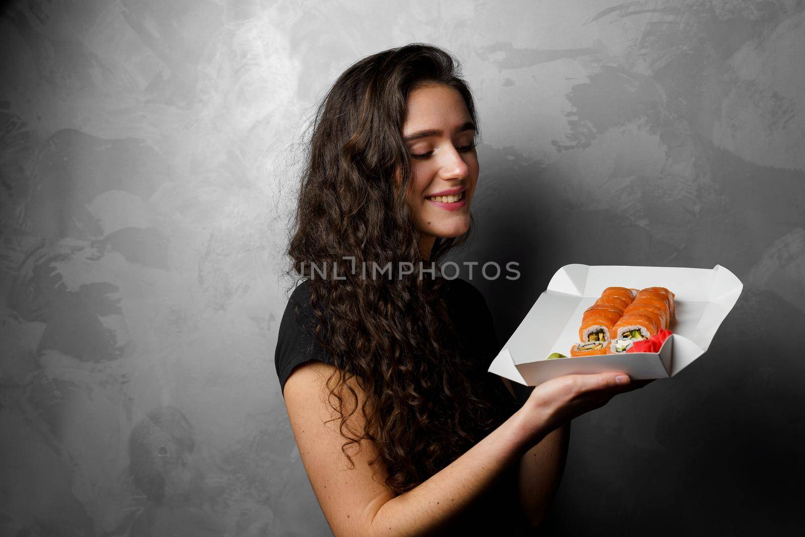 Surprised girl with sushi set philadelphia rolls in a paper box happy girl holding on a gray background. Food delivery. by Rabizo