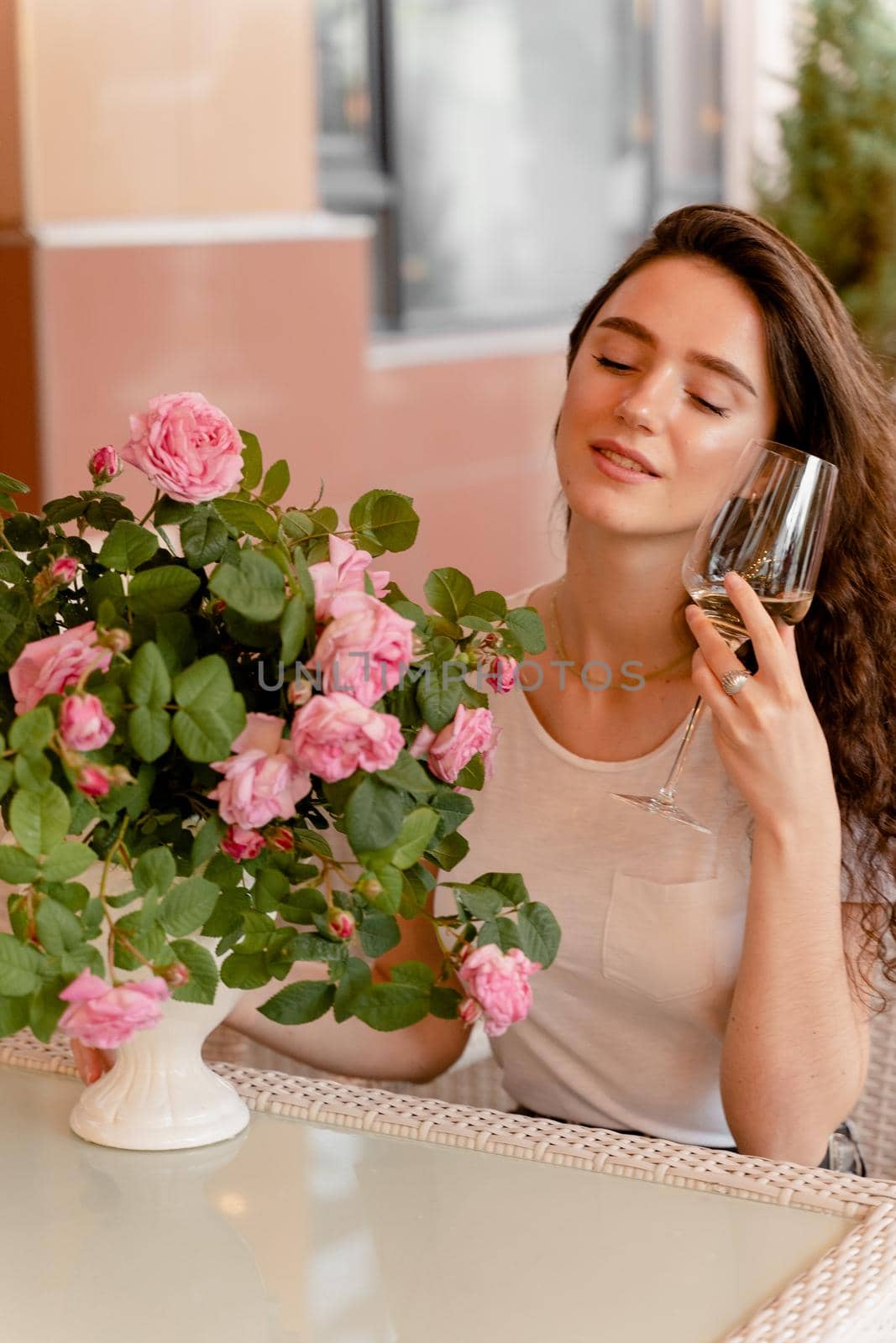 Dreaming woman with glass of alcoholic white wine in cafe in summer terrace and bouquet of roses on the table. by Rabizo