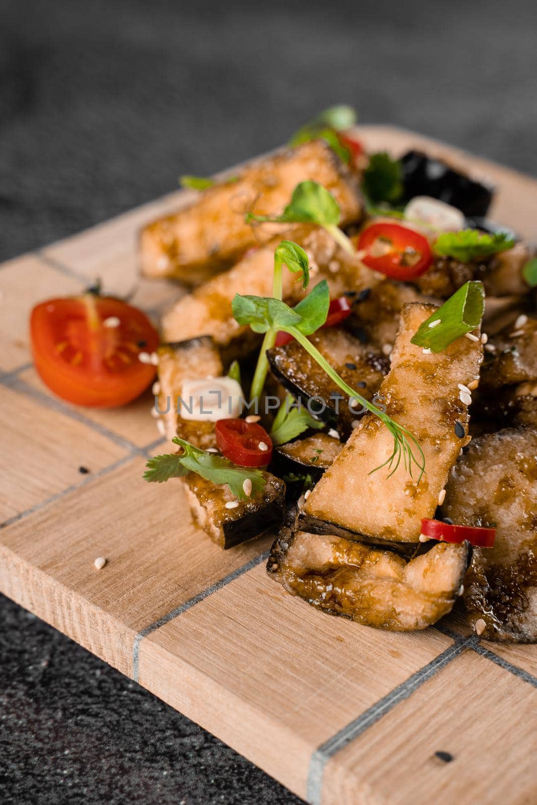 Fried eggplants on a wooden board, decorated with chili, garlic, cilantro, sesame seeds on a gray background. by Rabizo