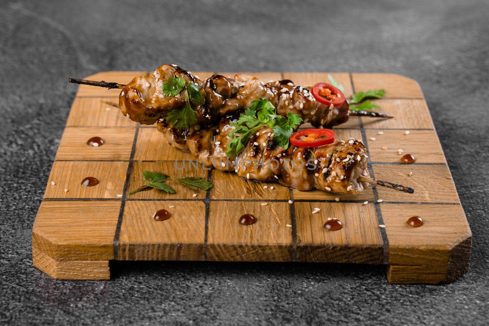 Meat on wooden skewers, garnished with cilantro, sesame, chili on square board on gray background