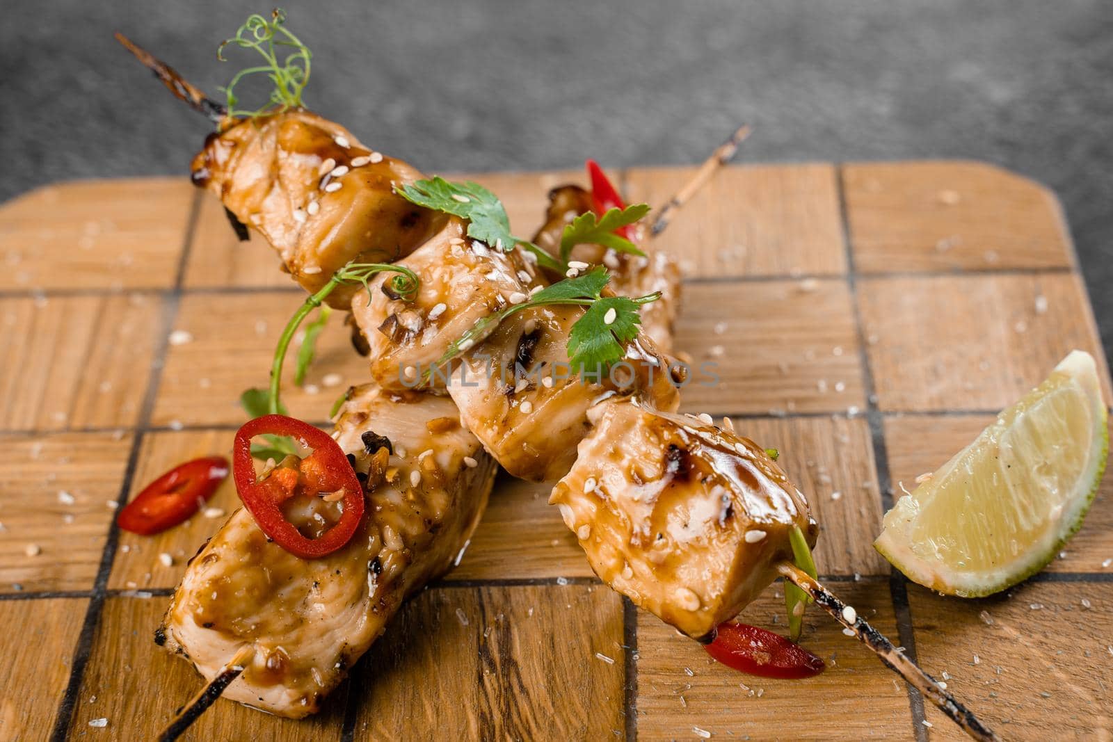 Meat on wooden skewers, garnished with cilantro, lime, sesame, chili on square board on gray background. by Rabizo