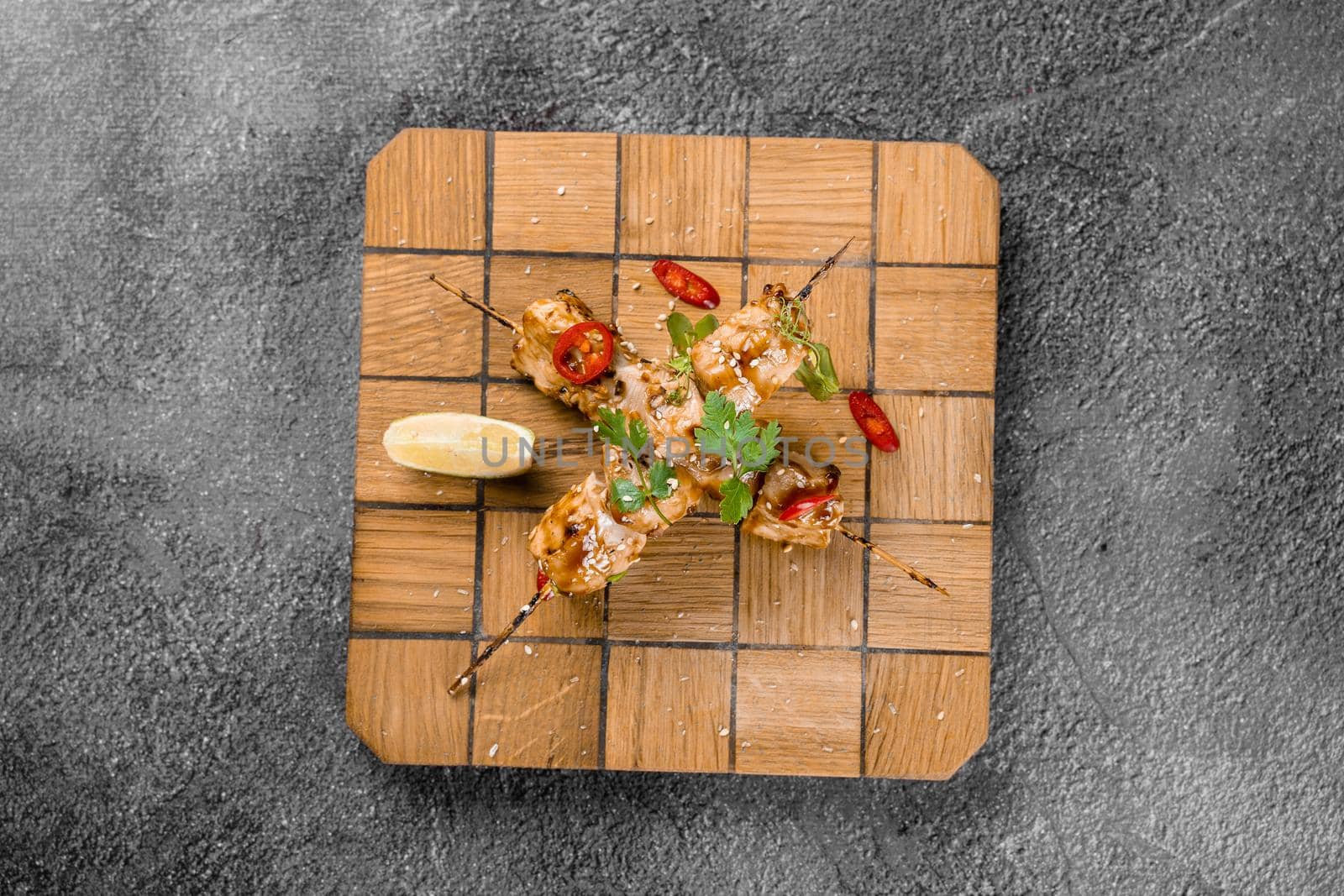 Meat on wooden skewers, garnished with cilantro, lime, sesame, chili on square board on gray background. by Rabizo