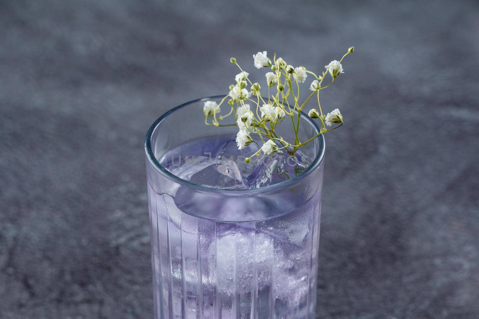 A cocktail in a tall glass with blue liqueur and ice, decorated with white flowers on gray background. by Rabizo