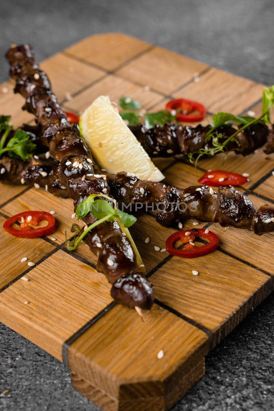 Grilled chicken hearts with lime on wooden skewer, garnished with chili, cilantro, sesame on gray background