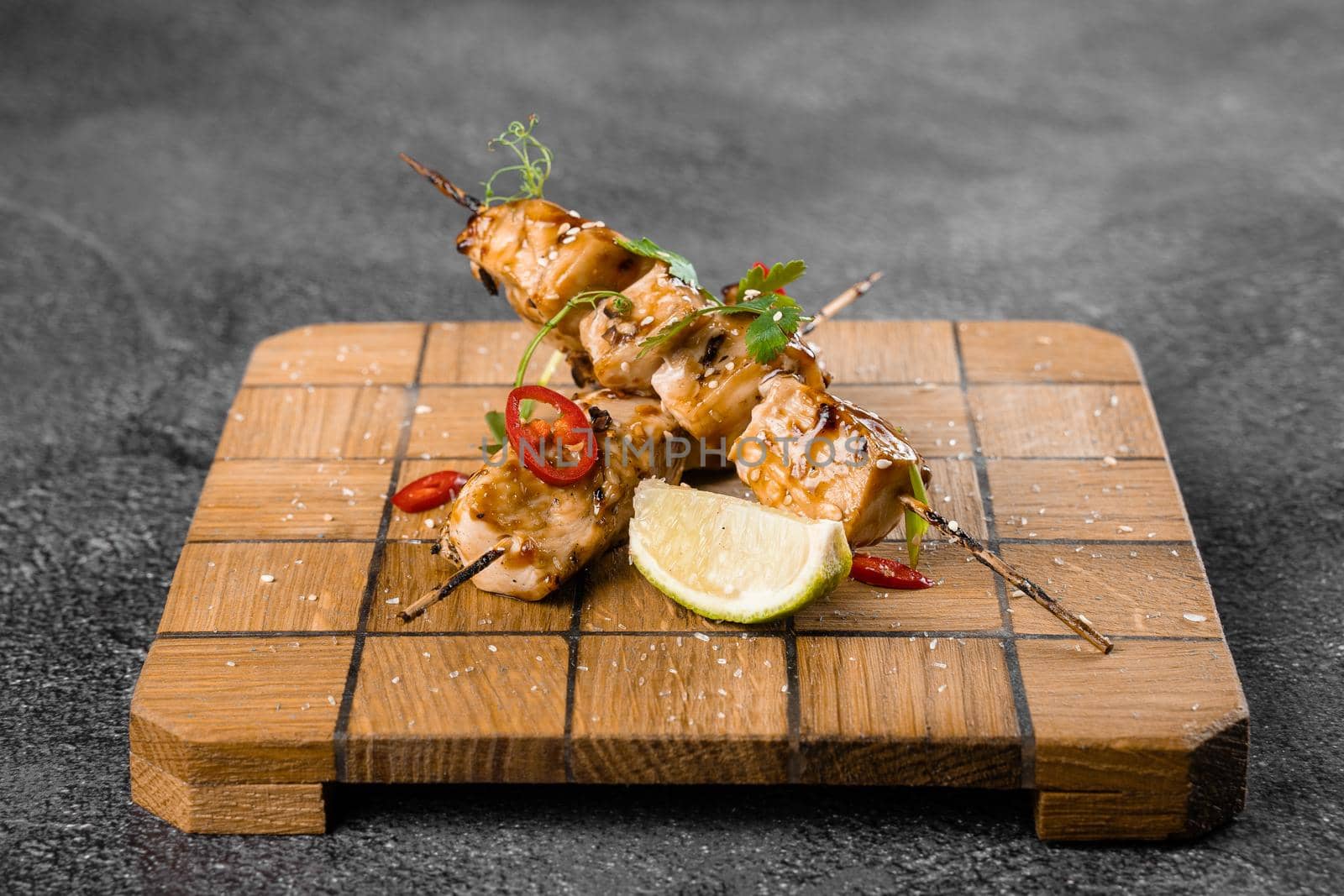 Meat on wooden skewers, garnished with cilantro, lime, sesame, chili on square board on gray background