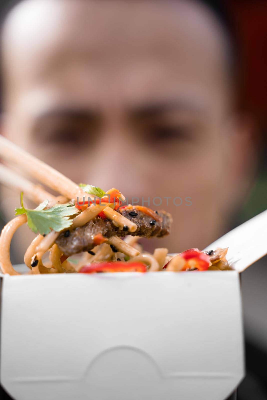 Chinese man holding spicy noodles with chopsticks. Wok in box in black food container. Fast food delivery service. Takeaway chinese street meal