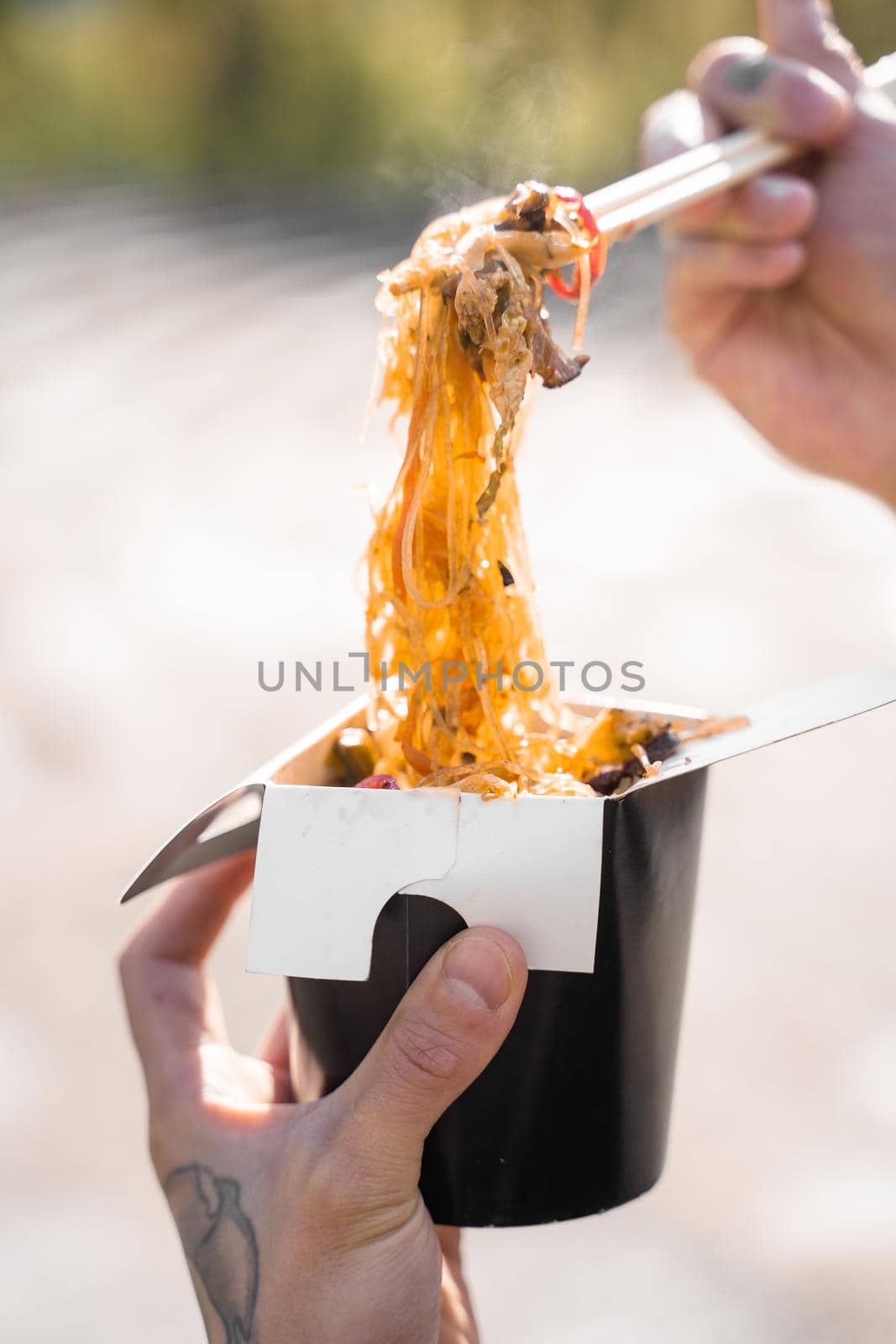 Wok in box in black food container. Holding rice noodles with chopsticks. Fast food delivery service. Takeaway chinese street meal. Eating udon by Rabizo