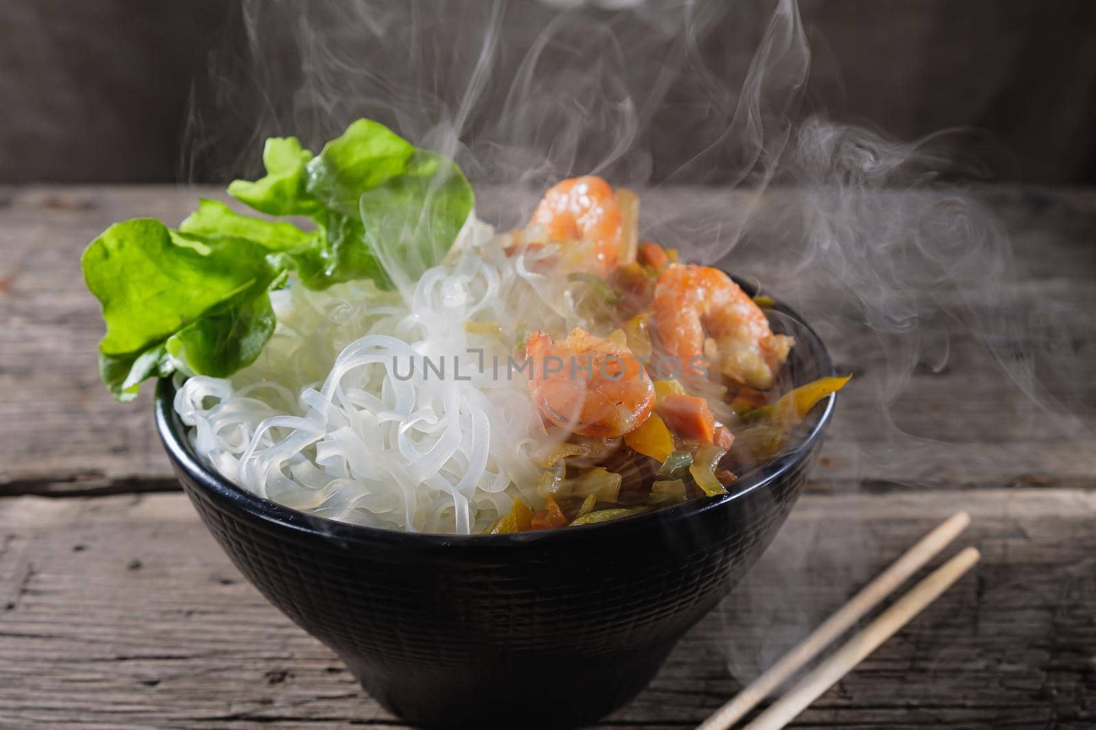 A plate of rice noodles stands on an old wooden table top. Rice noodles are most common in the cuisines of East, Southeast Asia and South Asia. The side dish consists of fried onions, carrots, paprika, shrimp. Copy space. Horizontal orientation