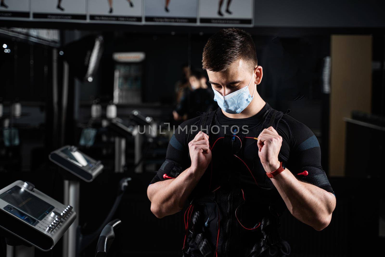 Man in EMS suit and medical mask in gym. Protection from coronavirus covid-19. Sport training in electrical muscle stimulation suit at quarantine period. by Rabizo