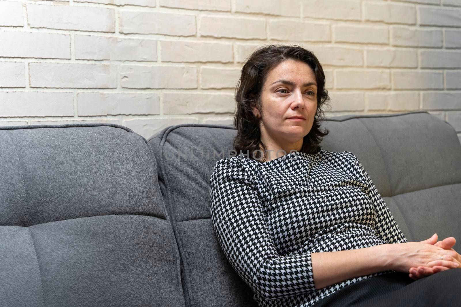 a middle-aged European woman with dark hair is sitting on a sofa against a light brick wall in a modern apartment. rest, thoughts