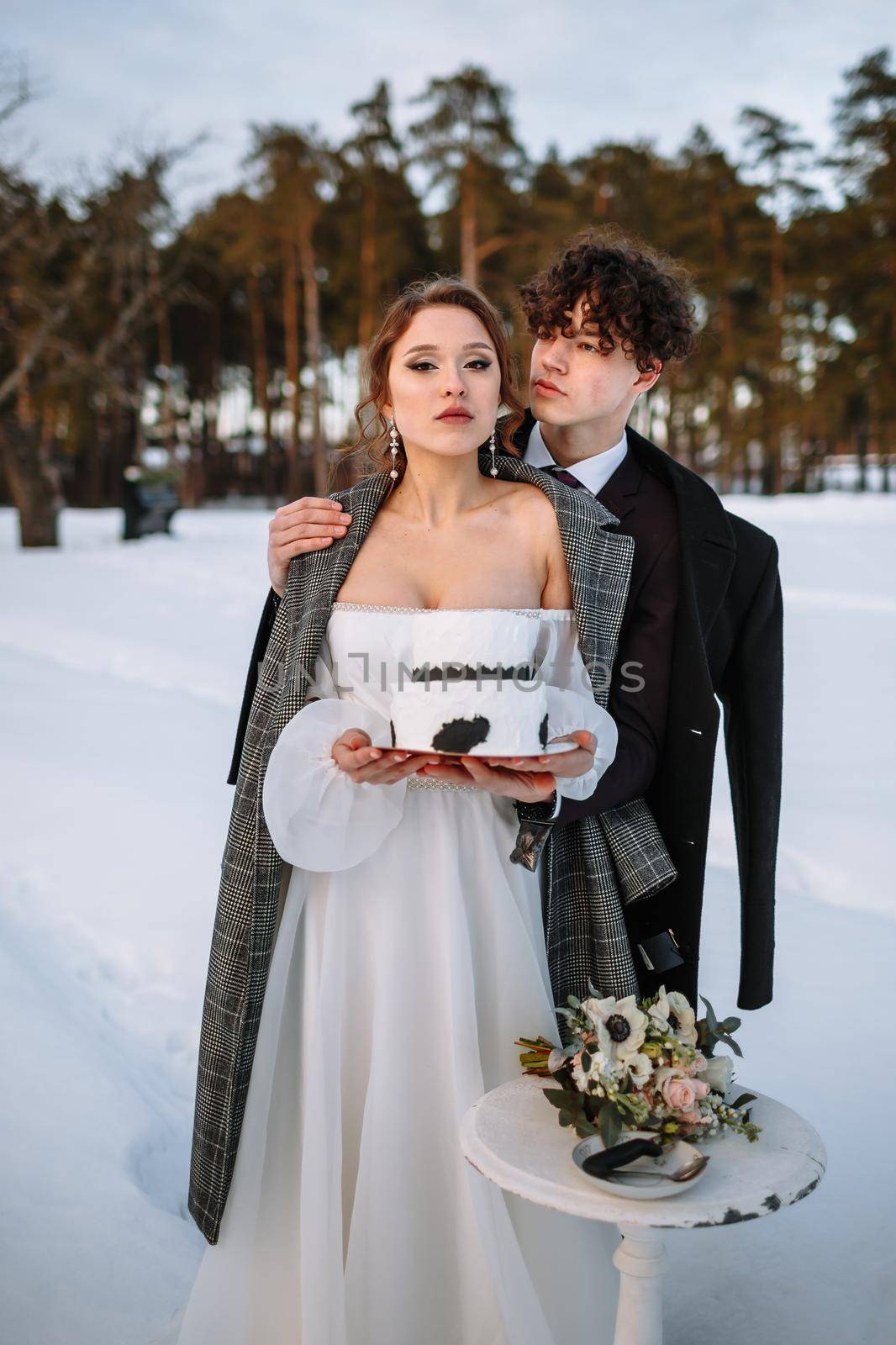 The bride and groom hold a wedding cake in their hands, standing in the snow in the forest.