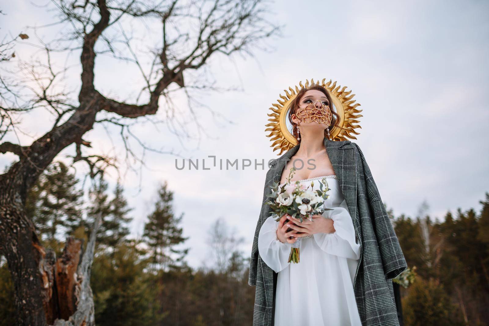 The bride is standing in a snow-covered forest. The bride in a fabulous protective mask. Above the bride is a halo in the form of sun rays