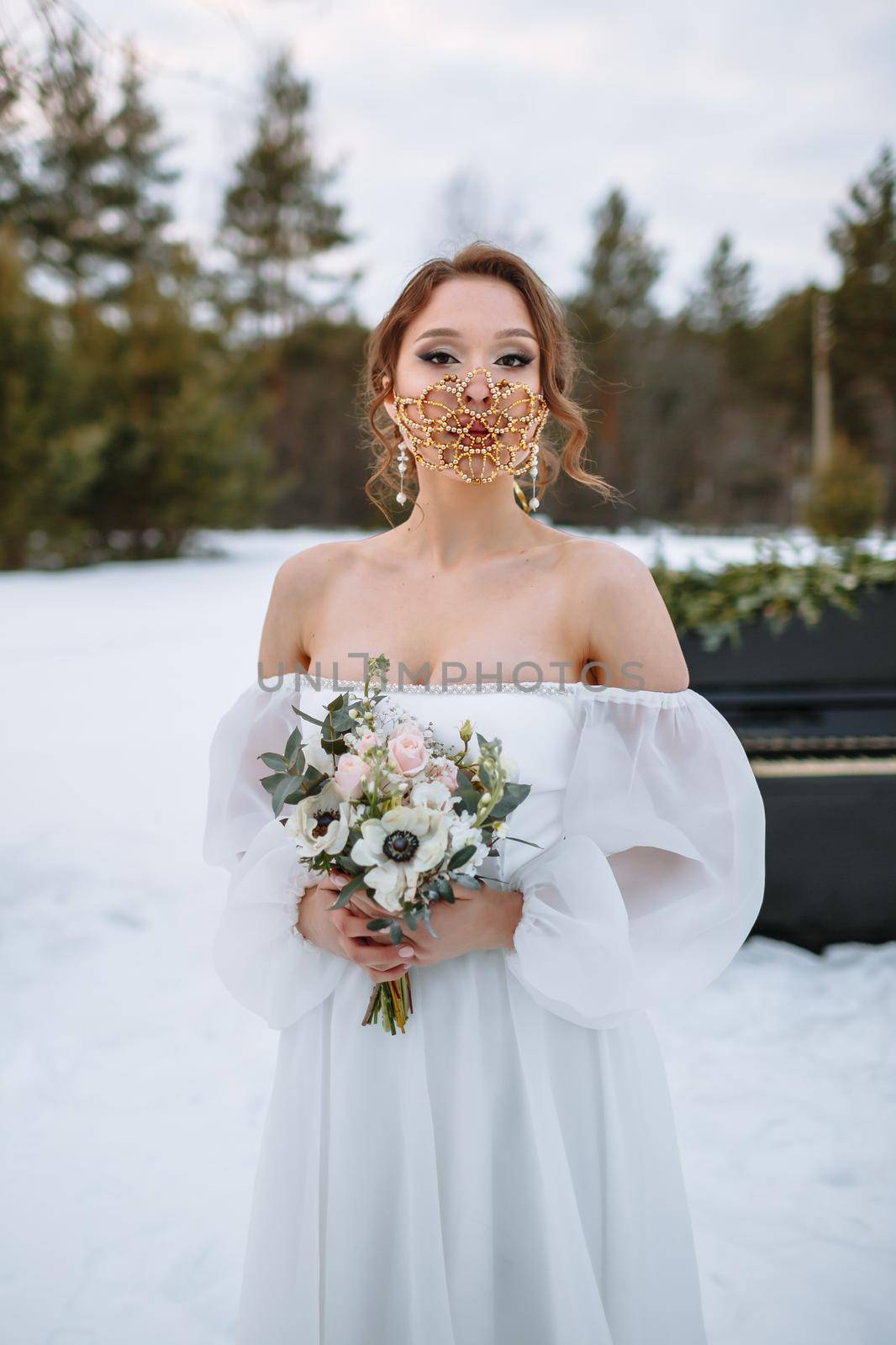 The bride is standing in a snow-covered forest. The bride is wearing a fabulous protective mask.