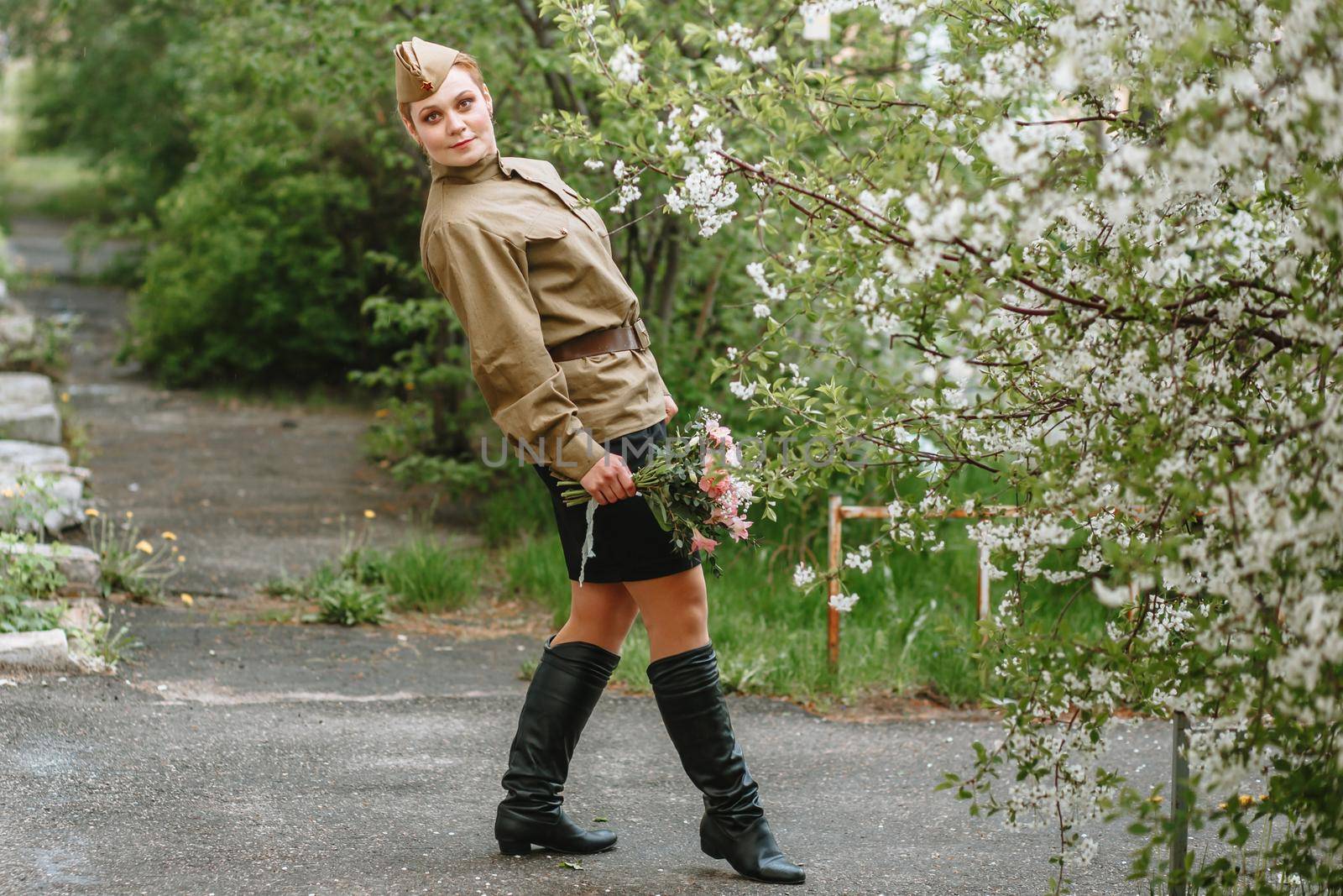 A girl in a Soviet military uniform next to a flowering tree. In the hands of a girl a beautiful bouquet