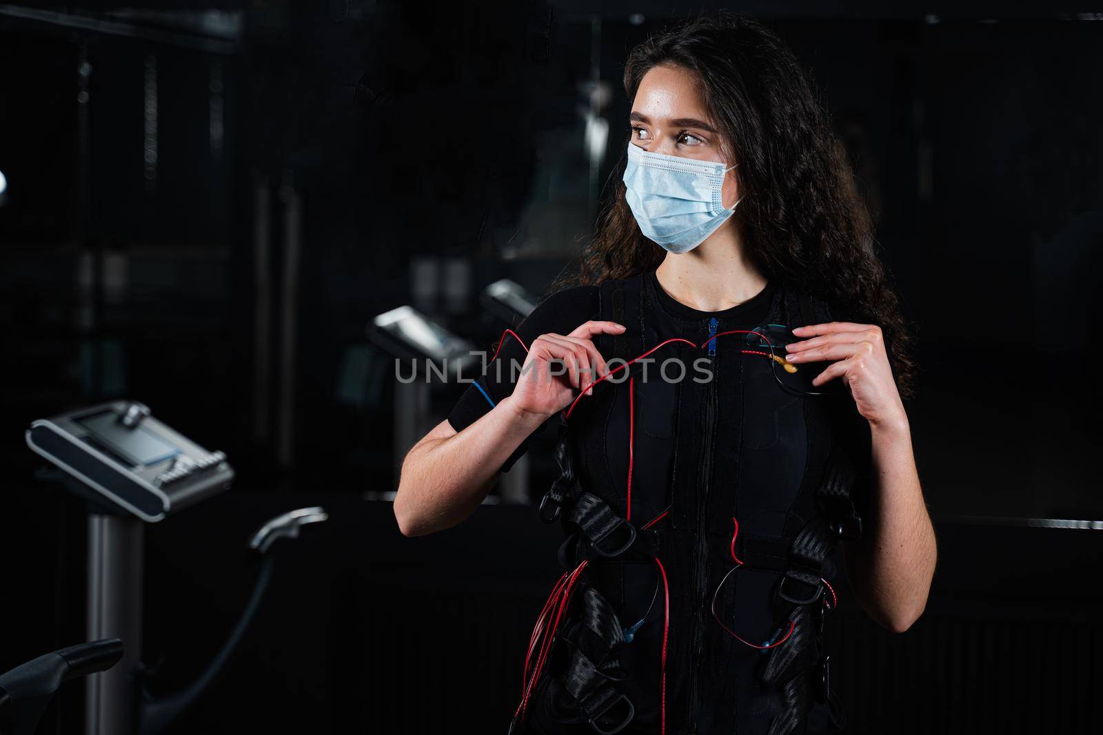 Girl in EMS suit and medical mask in gym. Protection from coronavirus covid-19. Sport training in electrical muscle stimulation suit at quarantine period