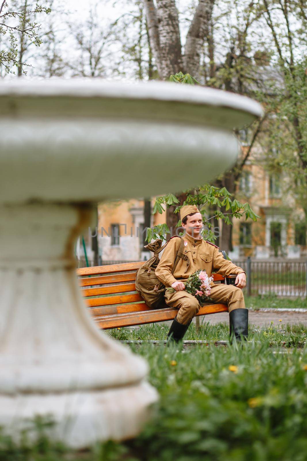 A soldier in a Soviet military uniform sits on a bench. In his hands a bouquet by deandy