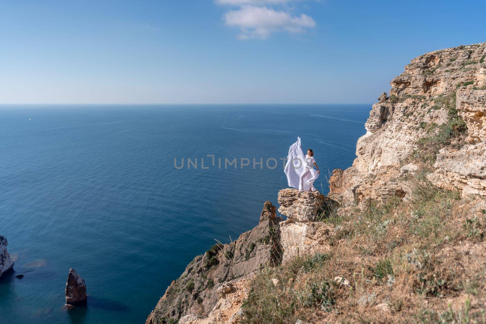 A beautiful young woman in a white light dress with long legs stands on the edge of a cliff above the sea waving a white long dress, against the background of the blue sky and the sea