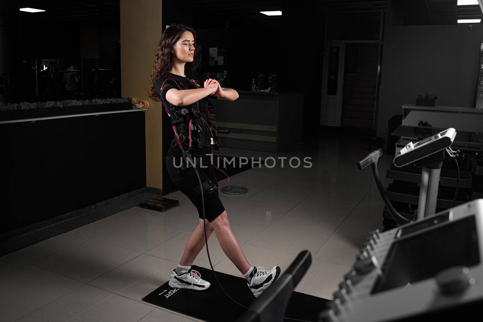 Girl in EMS suit in gym. Sport training in electrical muscle stimulation suit. Making physical exercises.