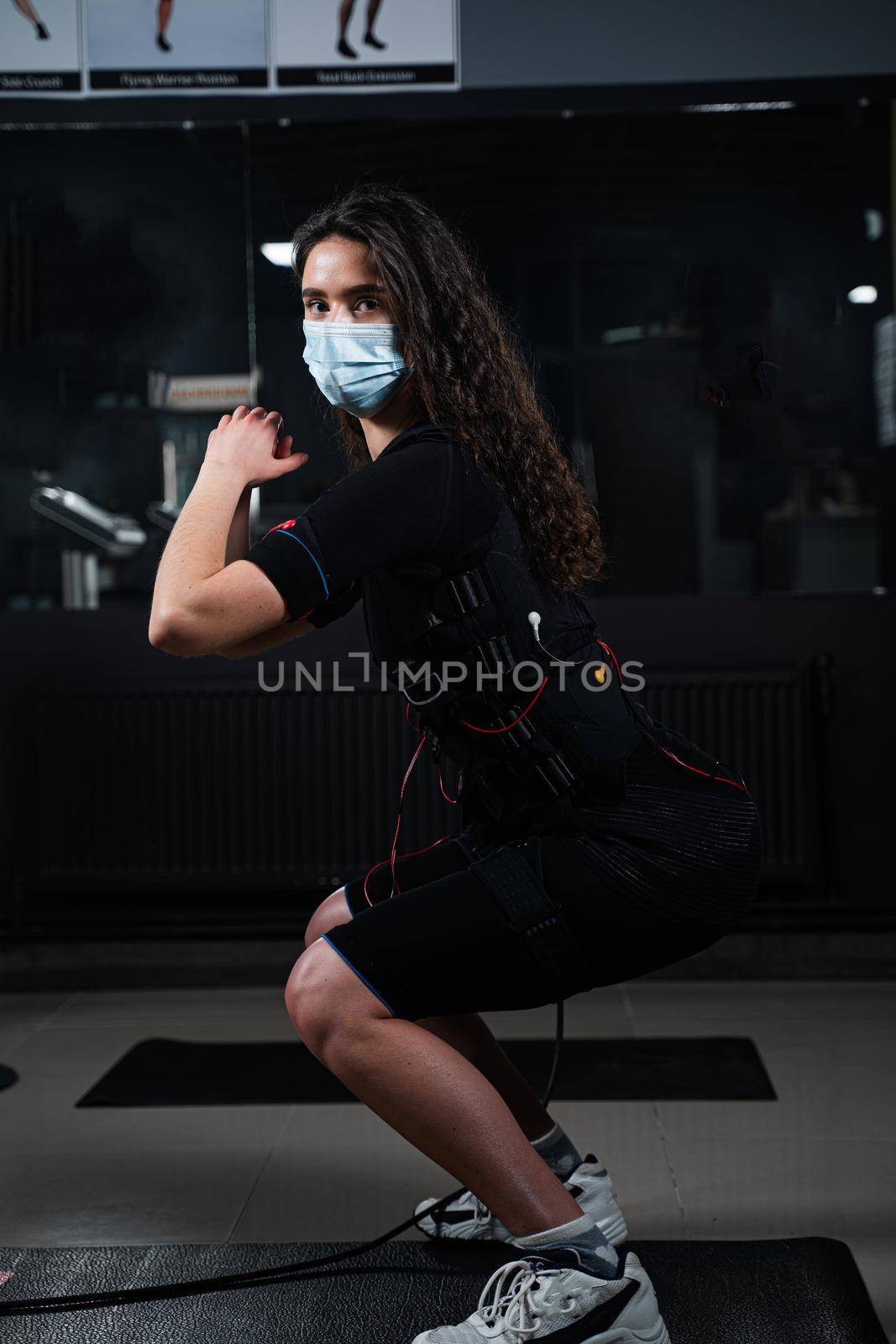 Girl in EMS suit and medical mask in gym. Protection from coronavirus covid-19. Sport training in electrical muscle stimulation suit at quarantine period. by Rabizo