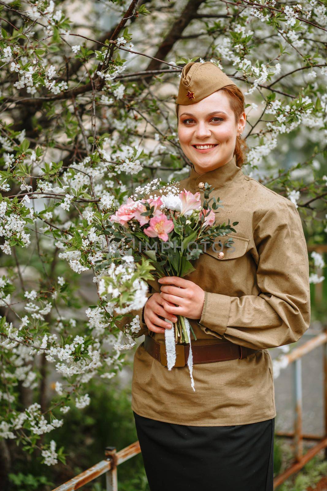 A girl in a Soviet military uniform next to a flowering tree. In the hands of a girl a beautiful bouquet by deandy