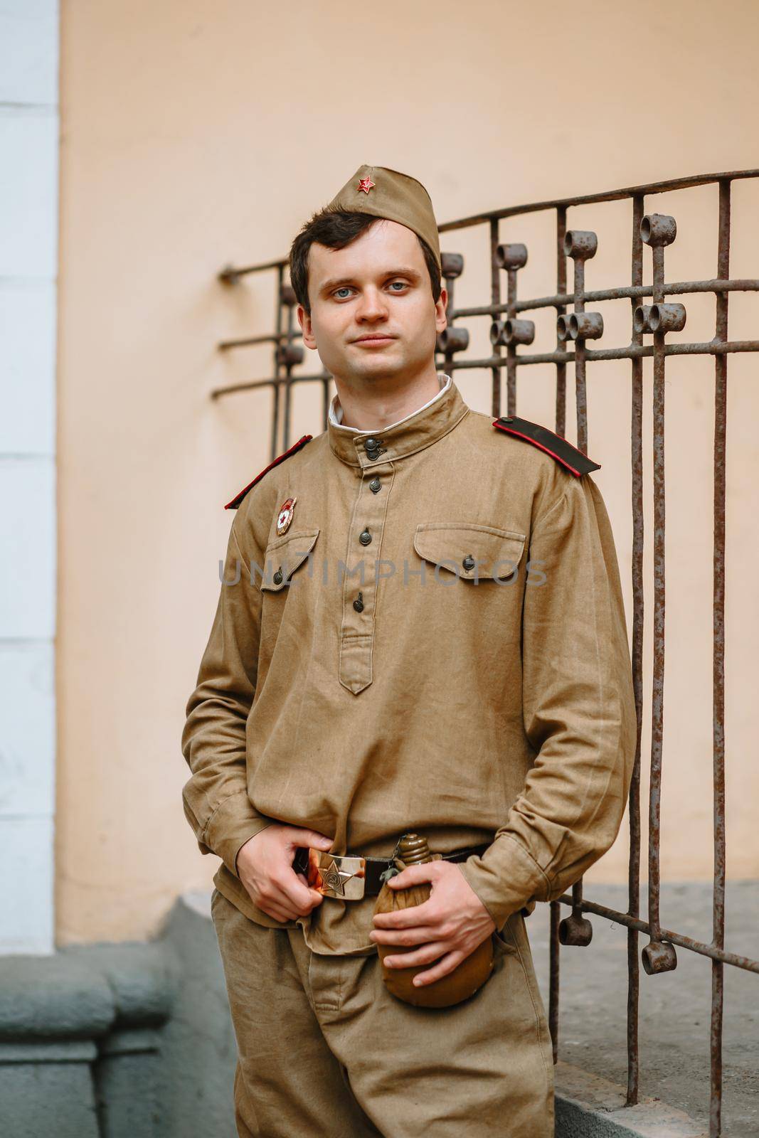 A man in a Soviet uniform from world war II. The soldier is standing against the yellow wall, holding his belt with his hands. Serious face