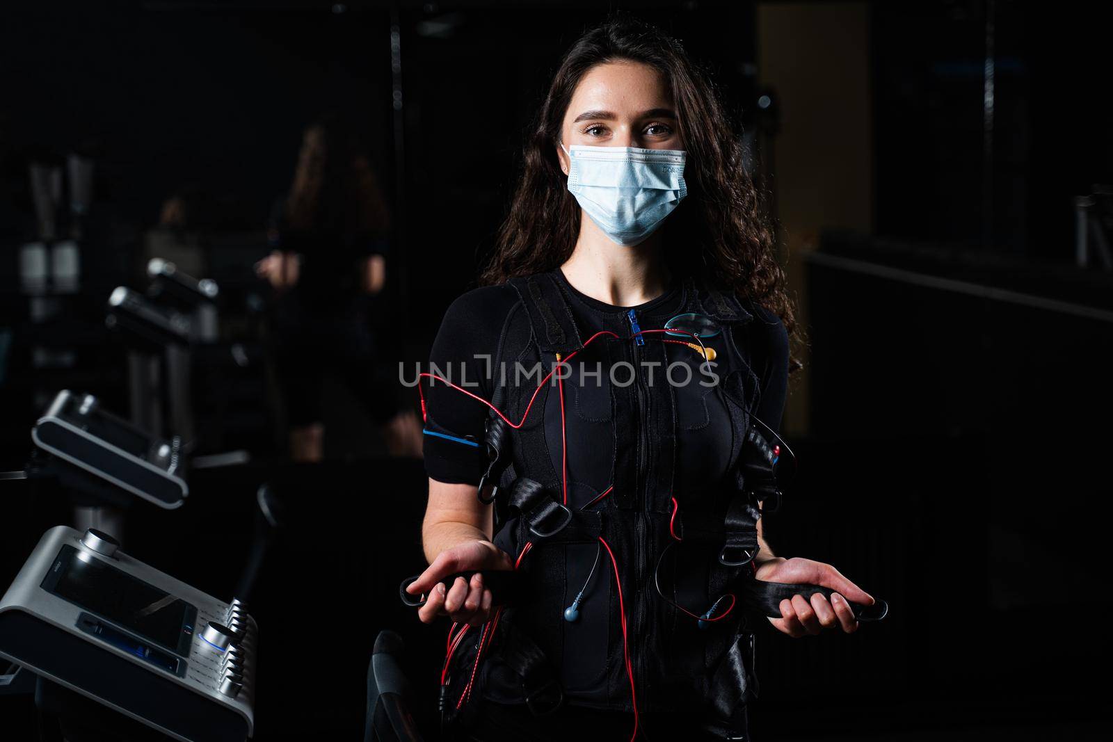 Girl in EMS suit and medical mask in gym. Protection from coronavirus covid-19. Sport training in electrical muscle stimulation suit at quarantine period