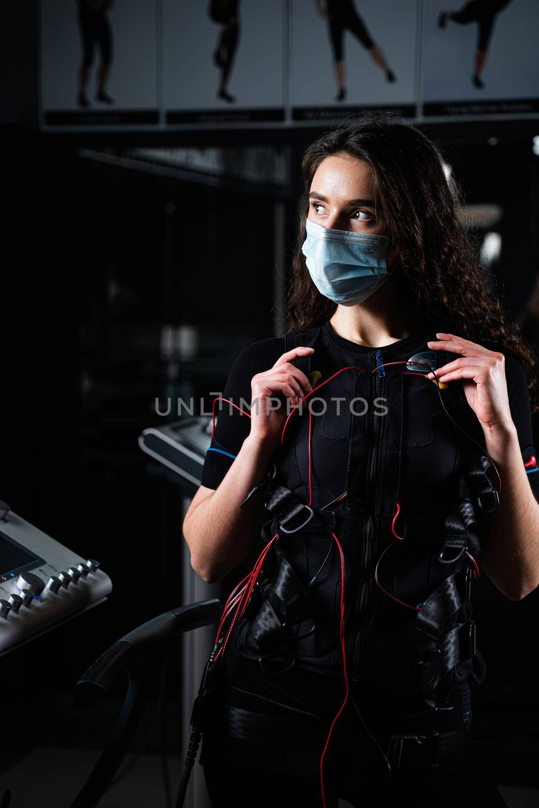 Girl in EMS suit and medical mask in gym. Protection from coronavirus covid-19. Sport training in electrical muscle stimulation suit at quarantine period. by Rabizo