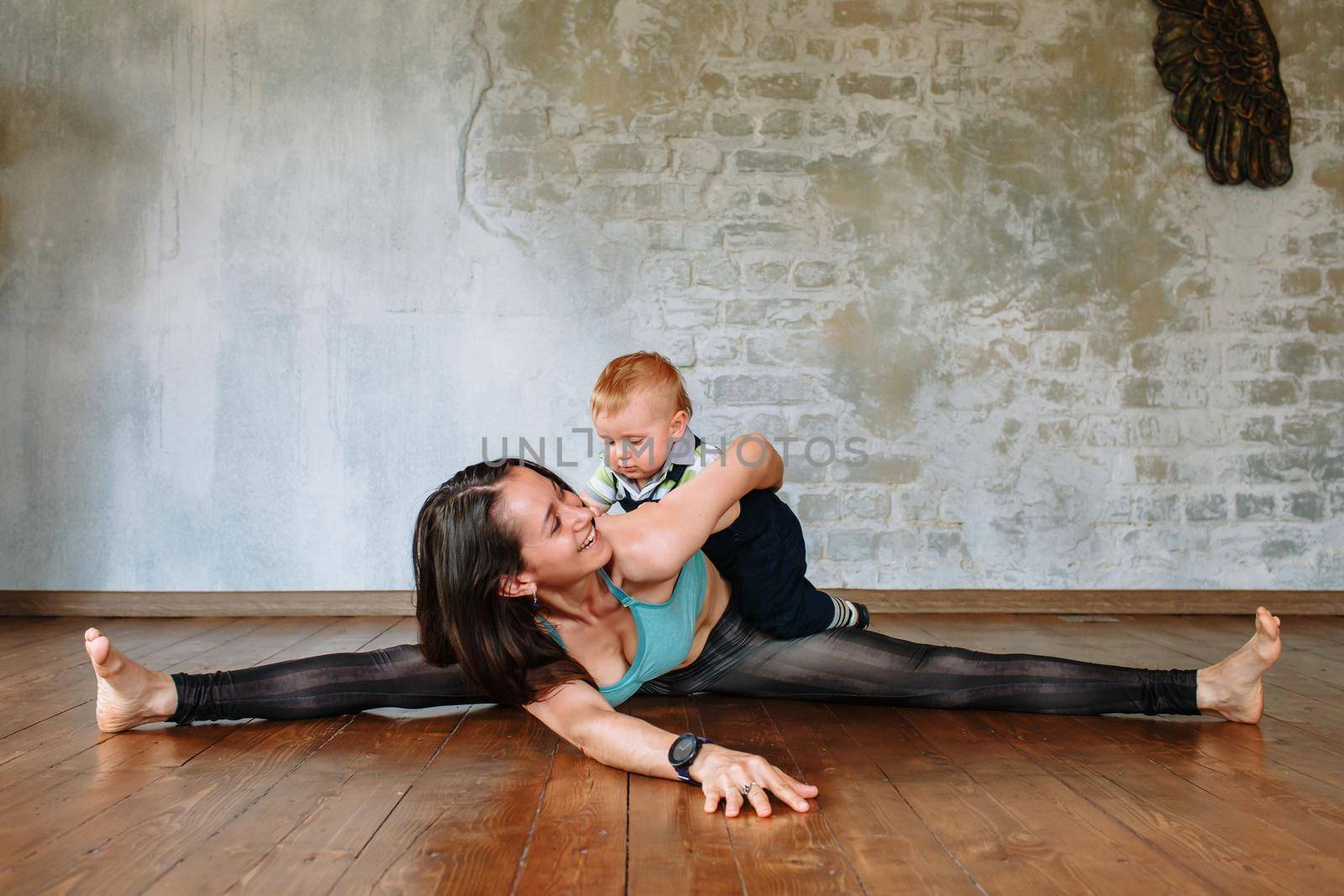 Yoga girl lies on the floor, on a wide splits and smiles. Her little son hugs her from behind by deandy