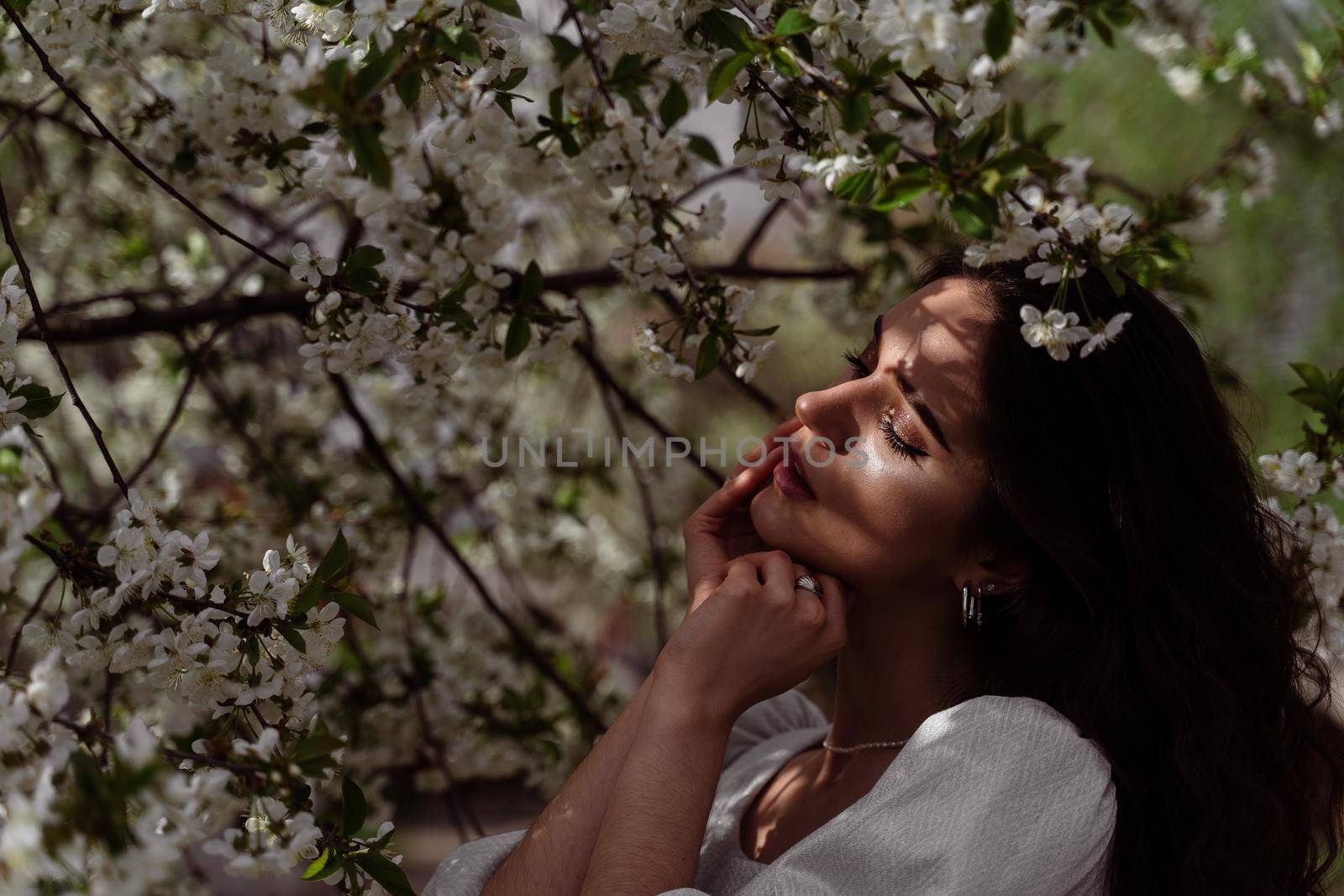 The sun shines on the girl's face near the flowering trees in the park. Portrait of attractive girl with curly hair in the garden