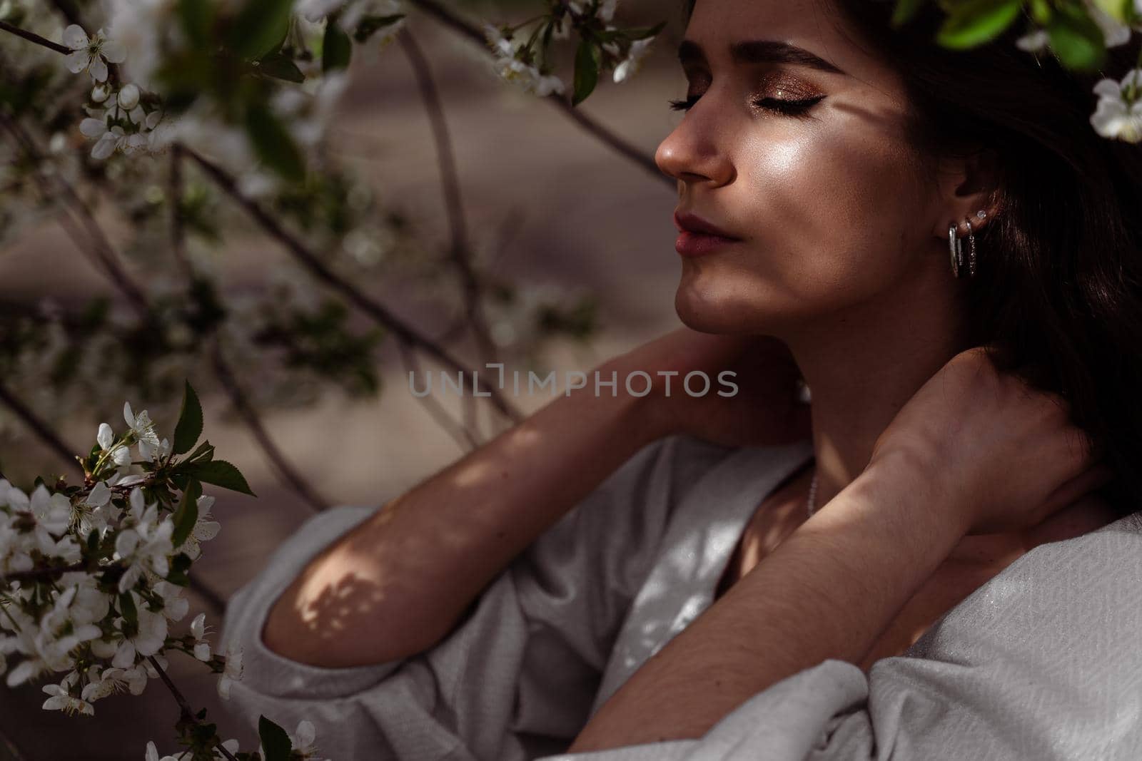 The sun shines on the girl's face near the flowering trees in the park. Portrait of attractive girl with curly hair in the garden