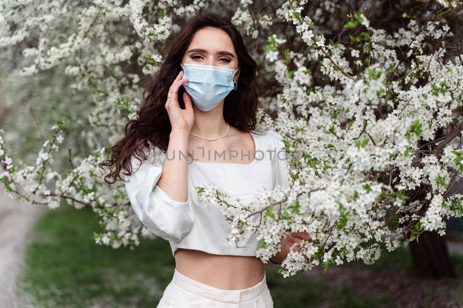 Girl in medical mask near white blooming trees in the park. Outdoor walking countryside at quarantine coronavirus covid-19 period. Spring lifestyle. by Rabizo