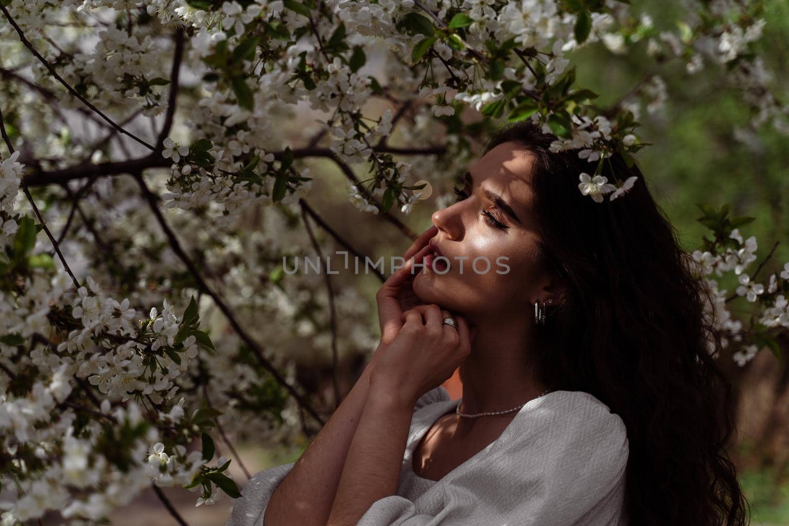 The sun shines on the girl's face near the flowering trees in the park. Portrait of attractive girl with curly hair in the garden