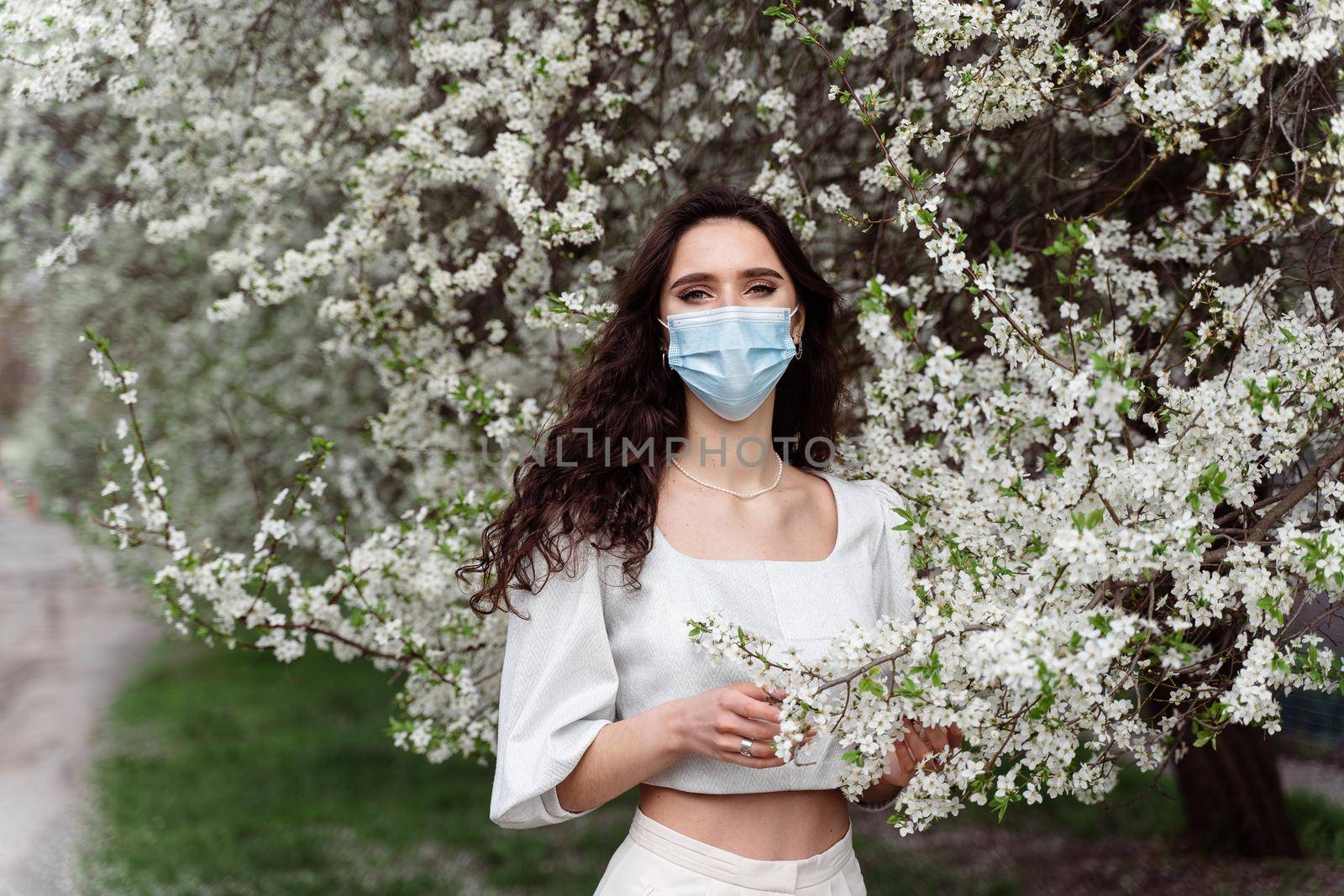 Girl in medical mask near white blooming trees in the park. Outdoor walking countryside at quarantine coronavirus covid-19 period. Spring lifestyle