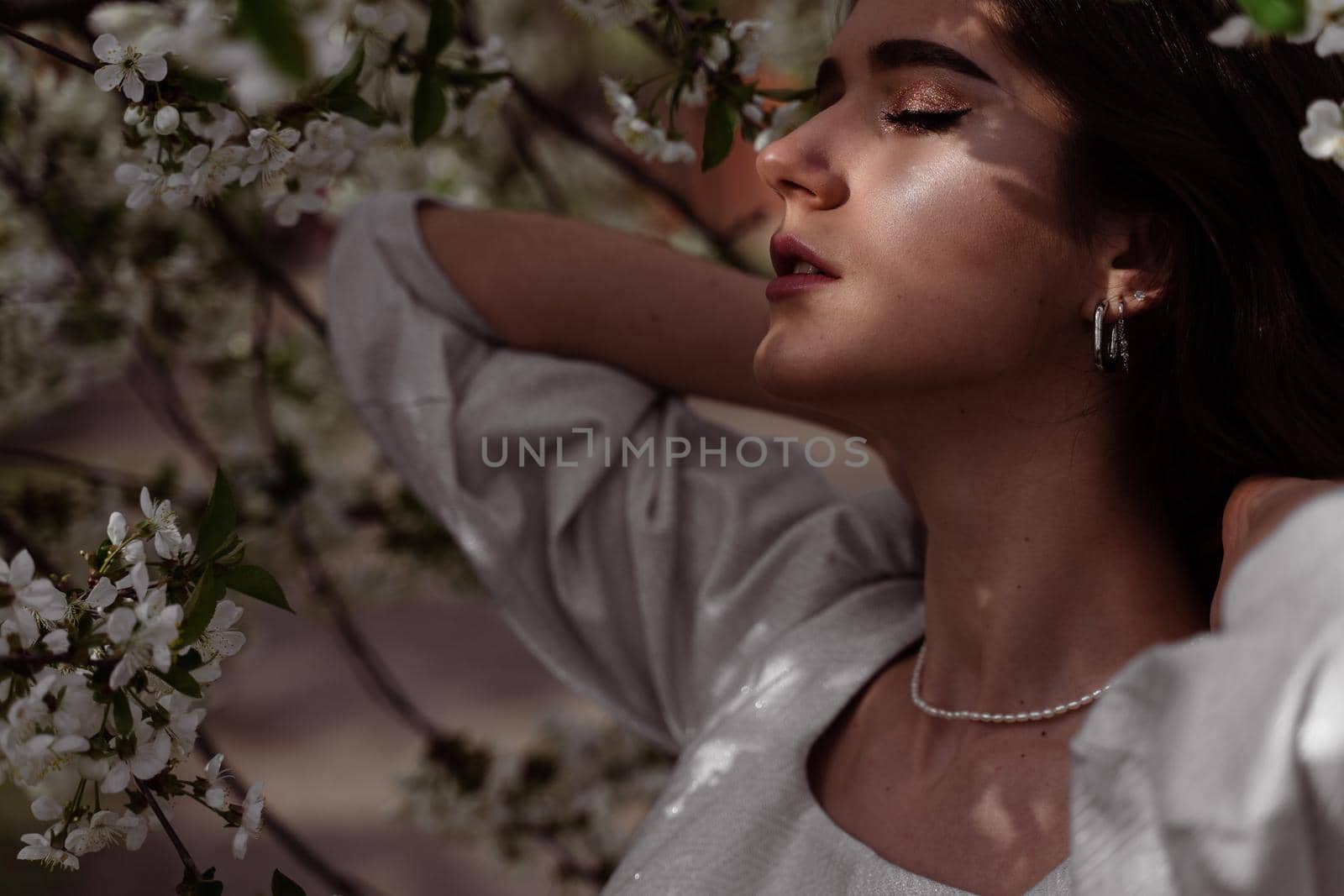 The sun shines on the girl's face near the flowering trees in the park. Portrait of attractive girl with curly hair in the garden