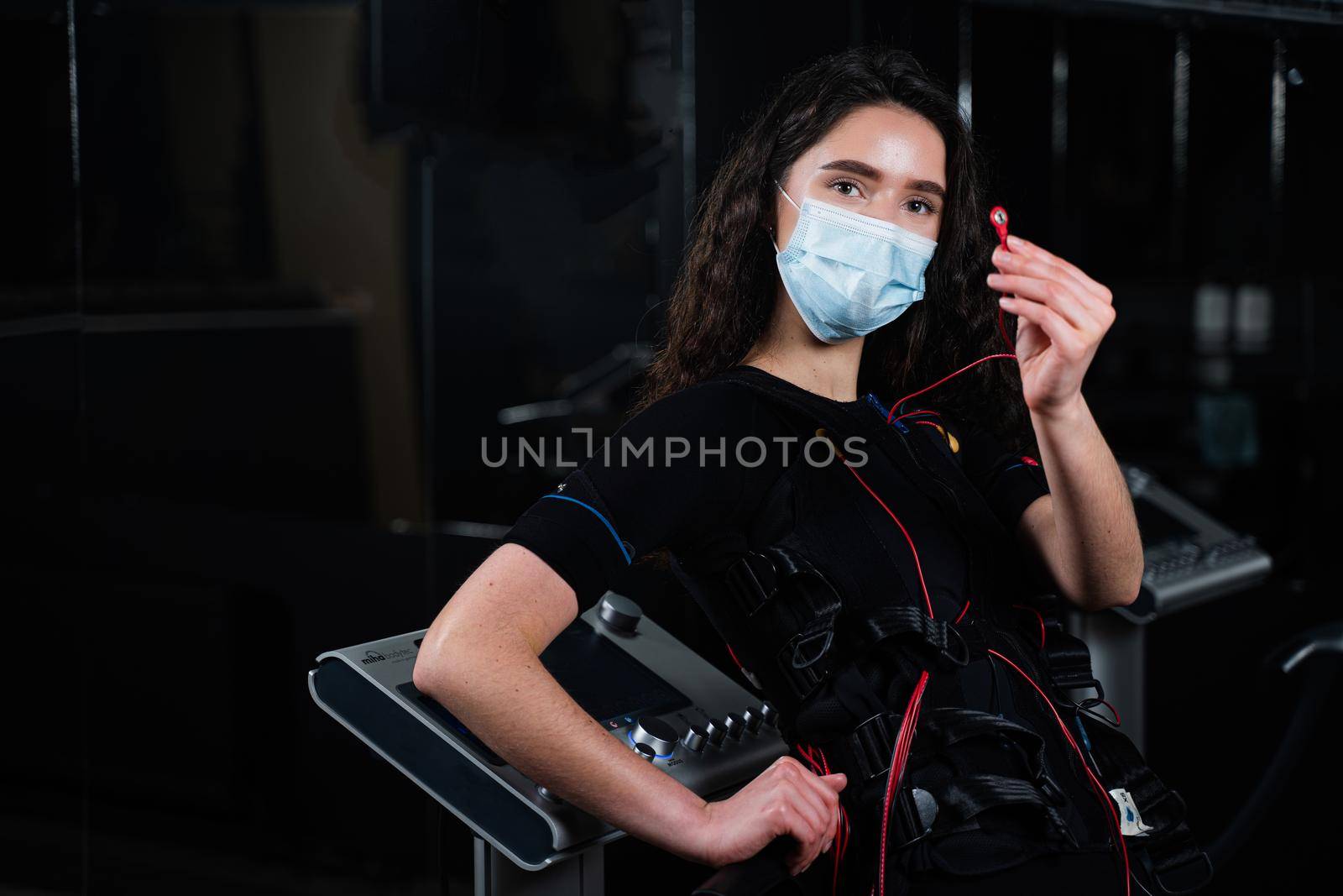 Girl in EMS suit and medical mask in gym. Protection from coronavirus covid-19. Sport training in electrical muscle stimulation suit at quarantine period