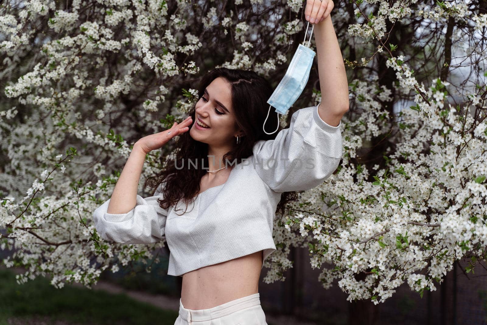 Girl holding medical mask after end of quarantine coronavirus covid-19 period. Model posing near white blooming trees in the garden