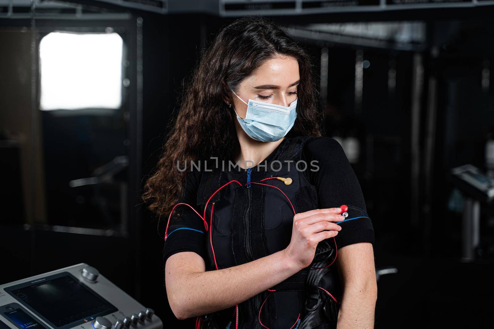 Girl in EMS suit and medical mask in gym. Protection from coronavirus covid-19. Sport training in electrical muscle stimulation suit at quarantine period