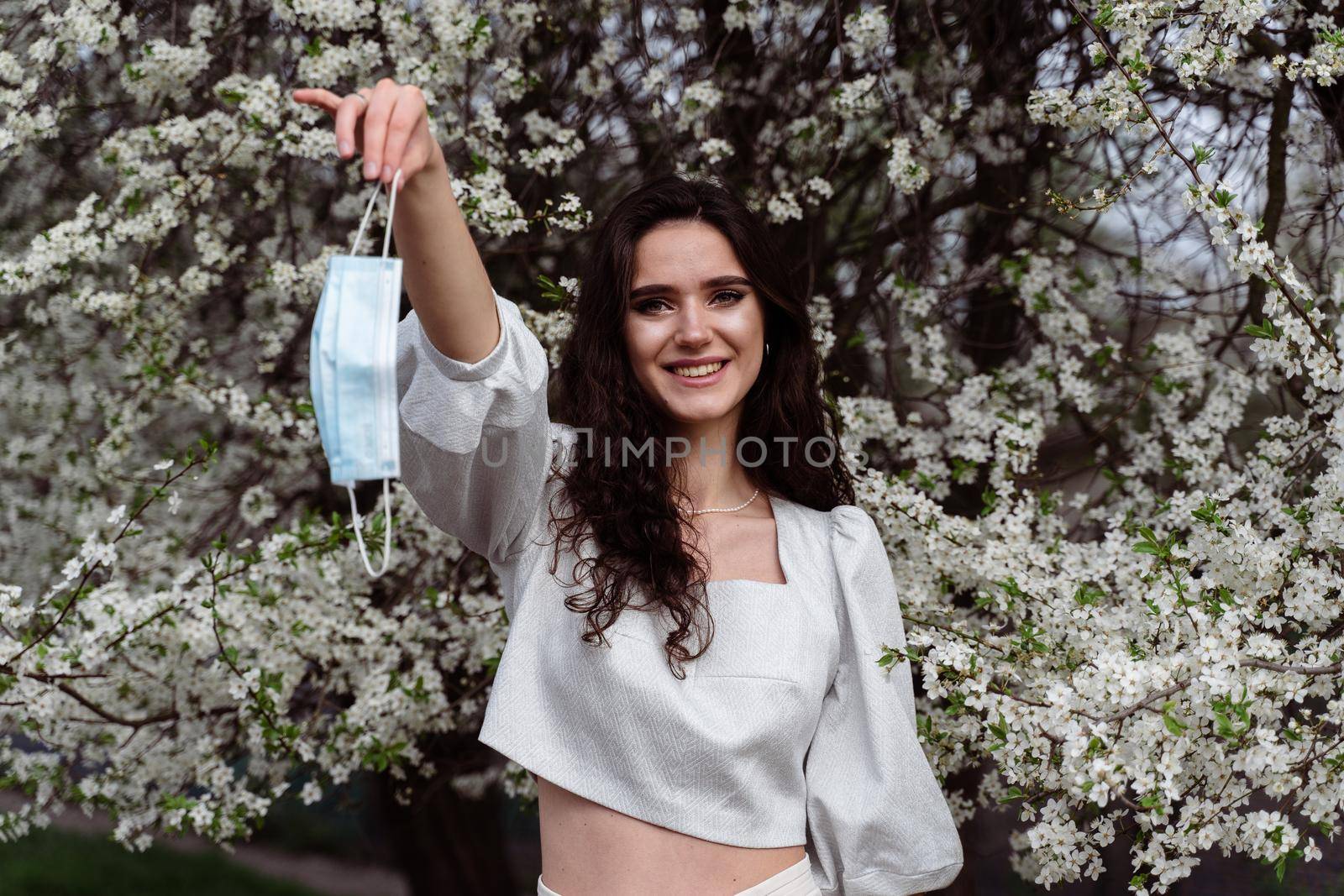 Girl holding medical mask after end of quarantine coronavirus covid-19 period. Model posing near white blooming trees in the garden