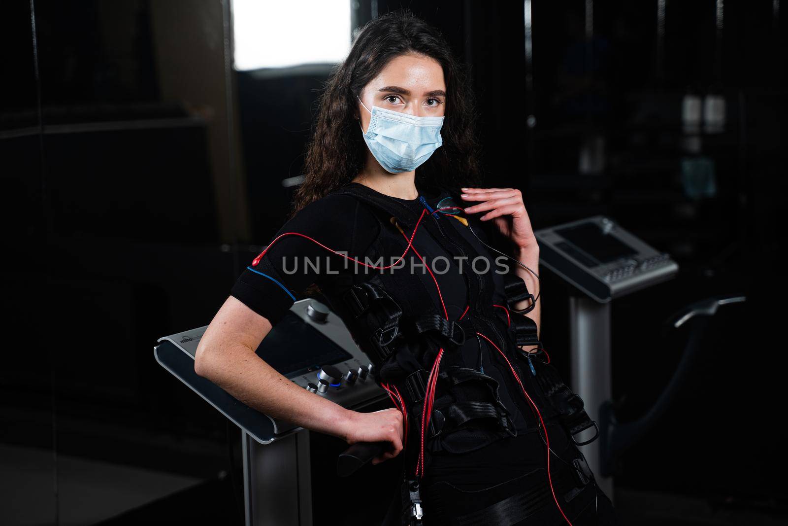 Girl in EMS suit and medical mask in gym. Protection from coronavirus covid-19. Sport training in electrical muscle stimulation suit at quarantine period. by Rabizo