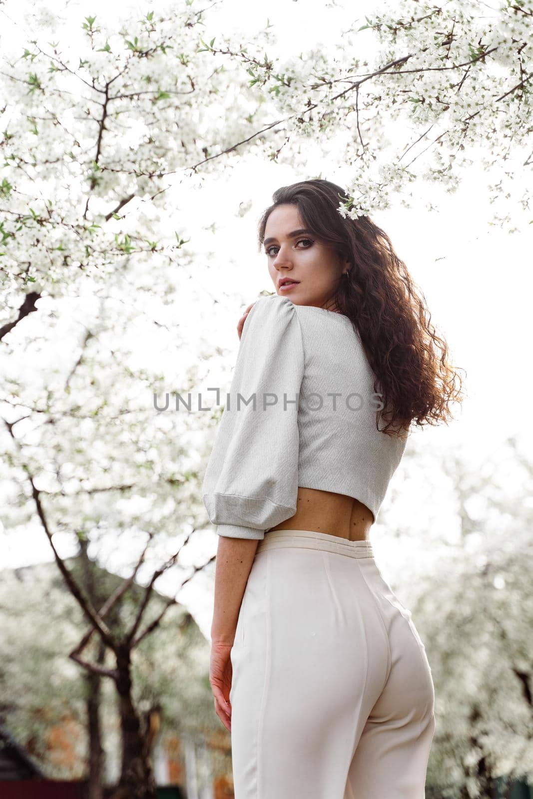 Spring lifestyle. Model posing near white blooming trees without mask outdoor countryside. Dreaming girl with curly hair