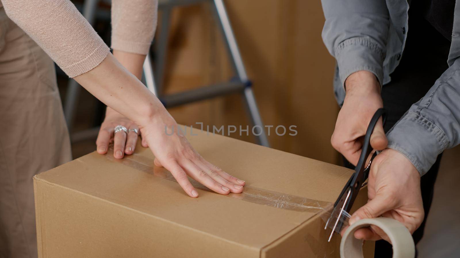Couple using adhesive tape to pack things in cardboard boxes, wrapping carton storage containers with sticky roller. Preparing packages for shipping transportation to move in. Close up.